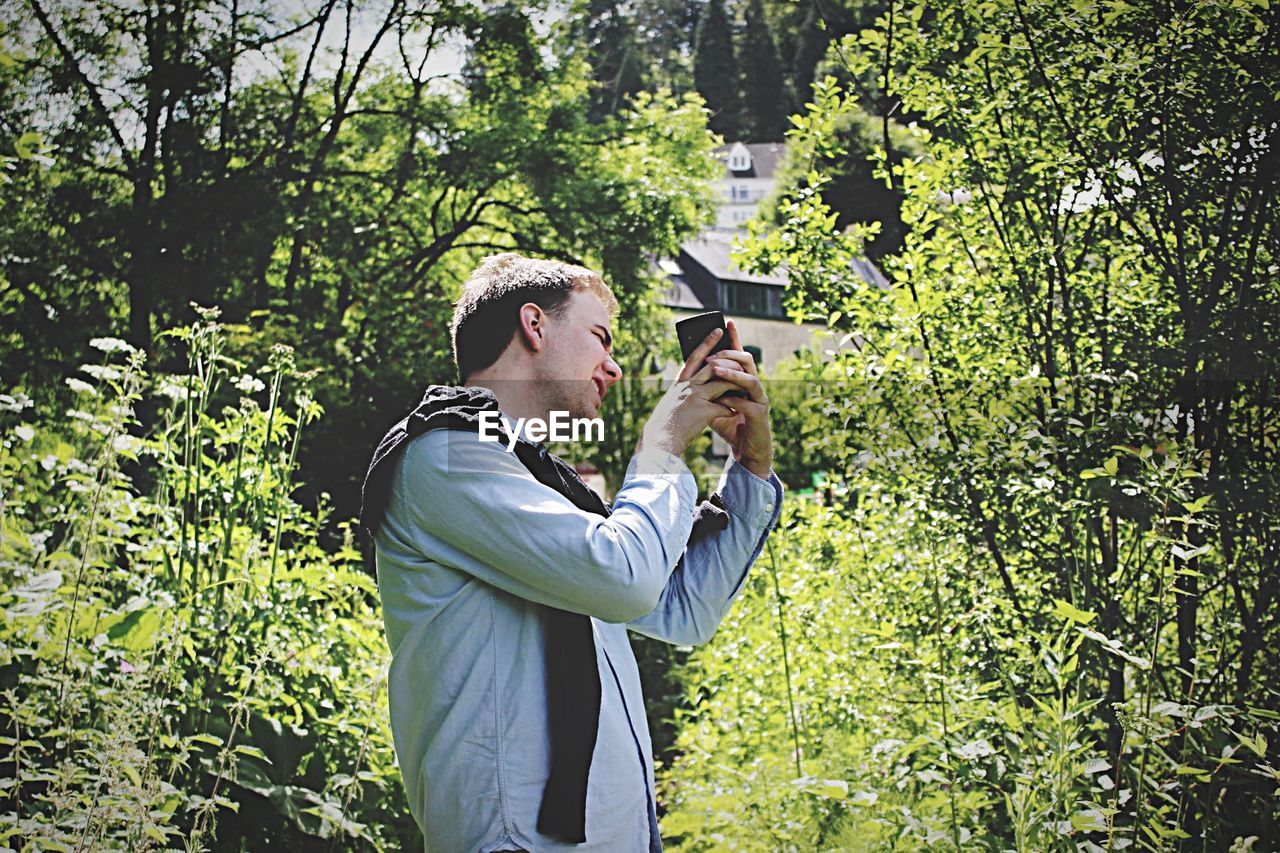 Side view of man photographing through mobile phone by plants