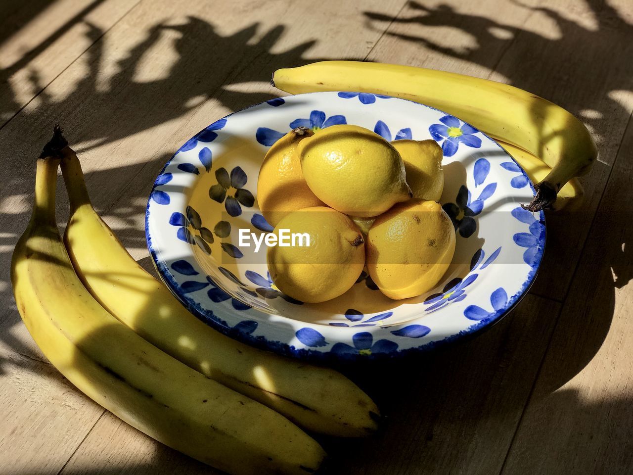 Lemons in blue flower patterned bowl surrounded by bananas on wooden table with sunlight , shadows.