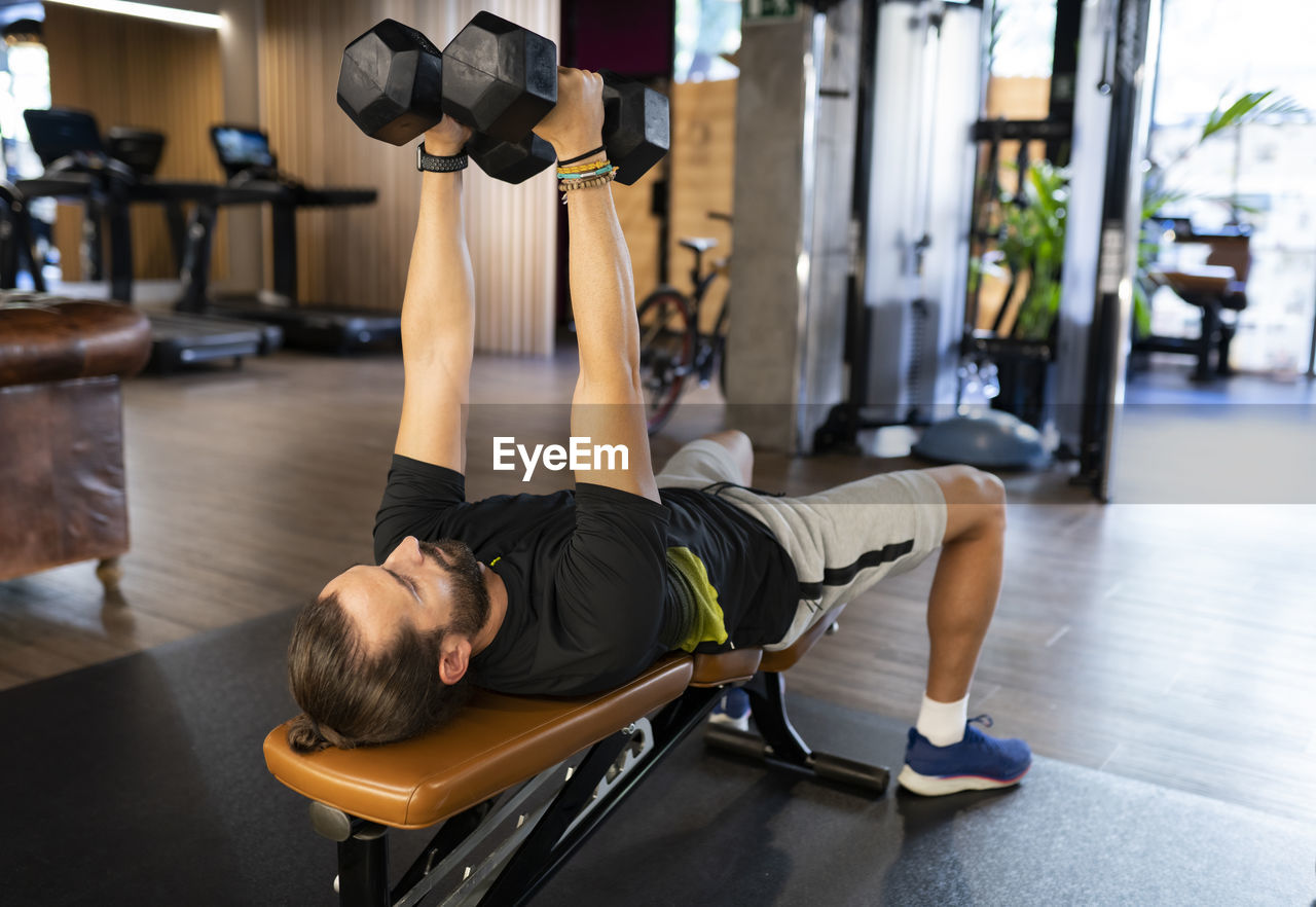 Bearded adult sportsman lying on bench and exercising with dumbbells during weightlifting training in modern gym