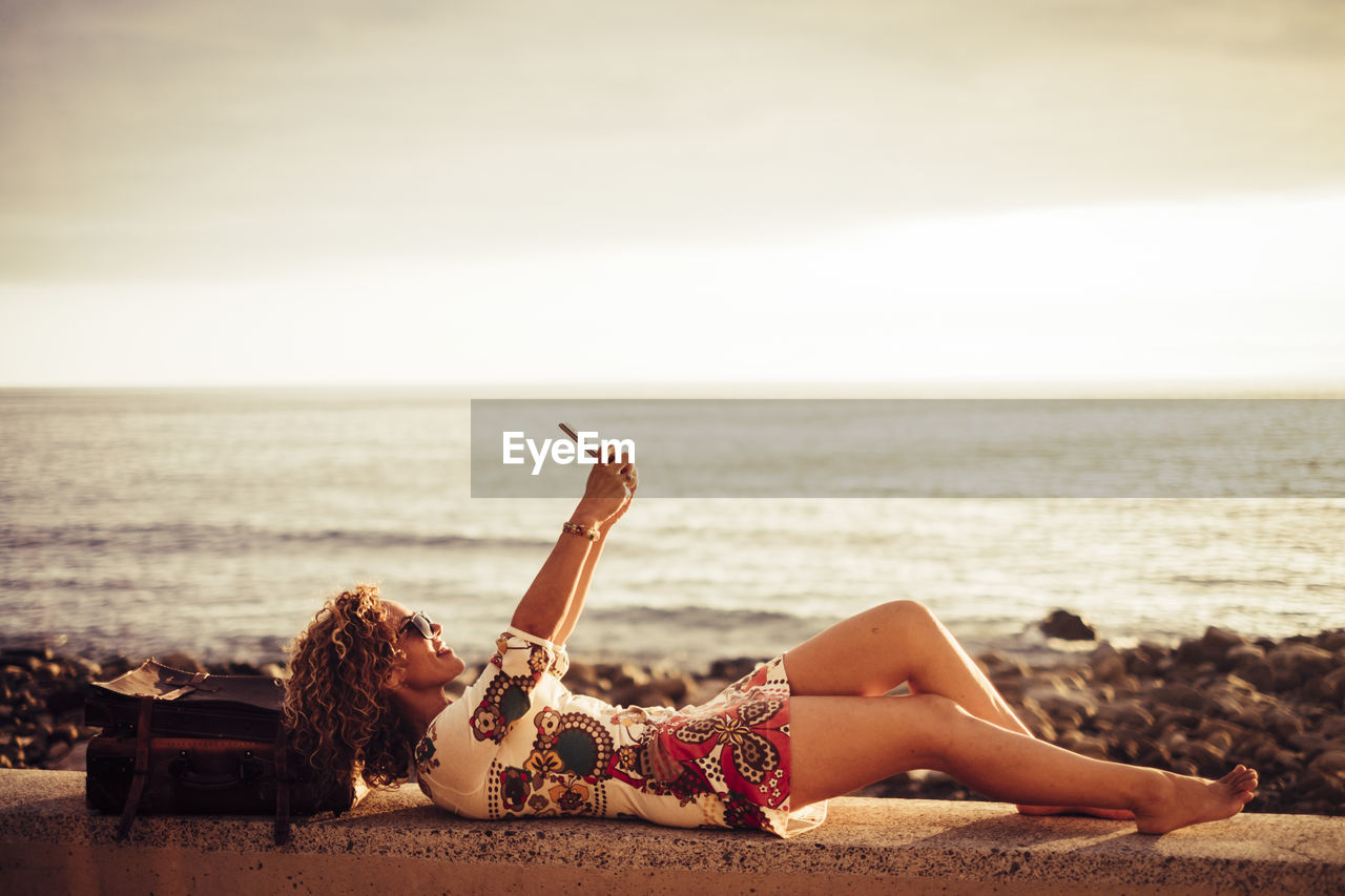 Side view of woman taking selfie while lying on retaining wall at beach against sky during sunset