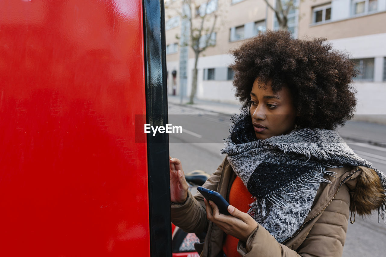 Afro woman with smart phone buying ticket from machine during winter