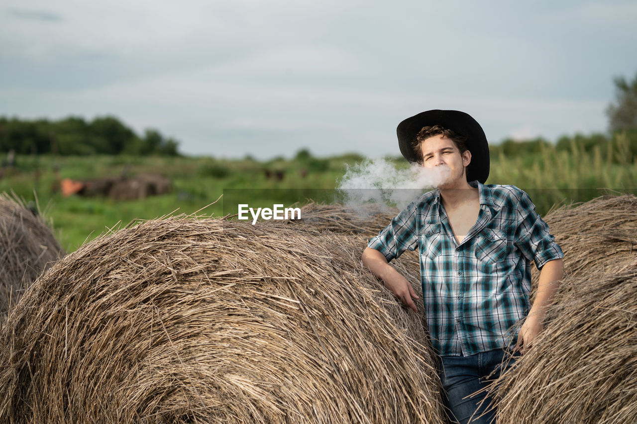 Portrait of a handsome young man in a cowboy hat vaping an electronic cigarette near the haystack.