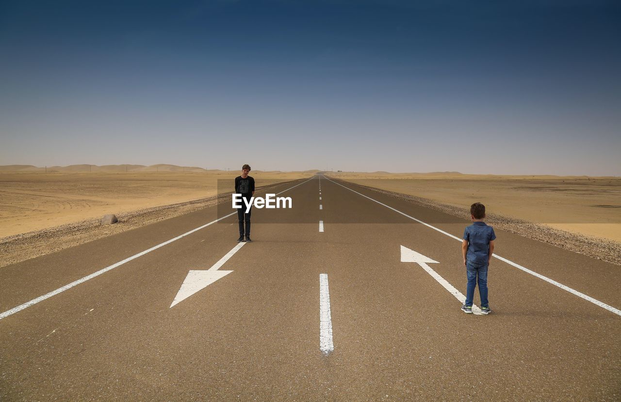 Brothers standing on road arrow symbols along landscape against clear blue sky