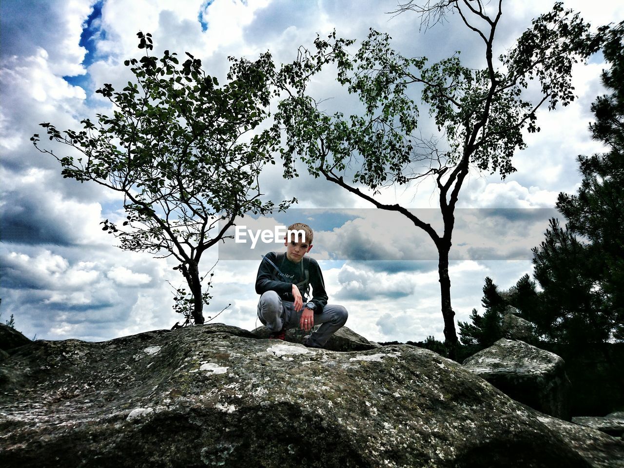 Low angle portrait of boy crouching on rock against cloudy sky