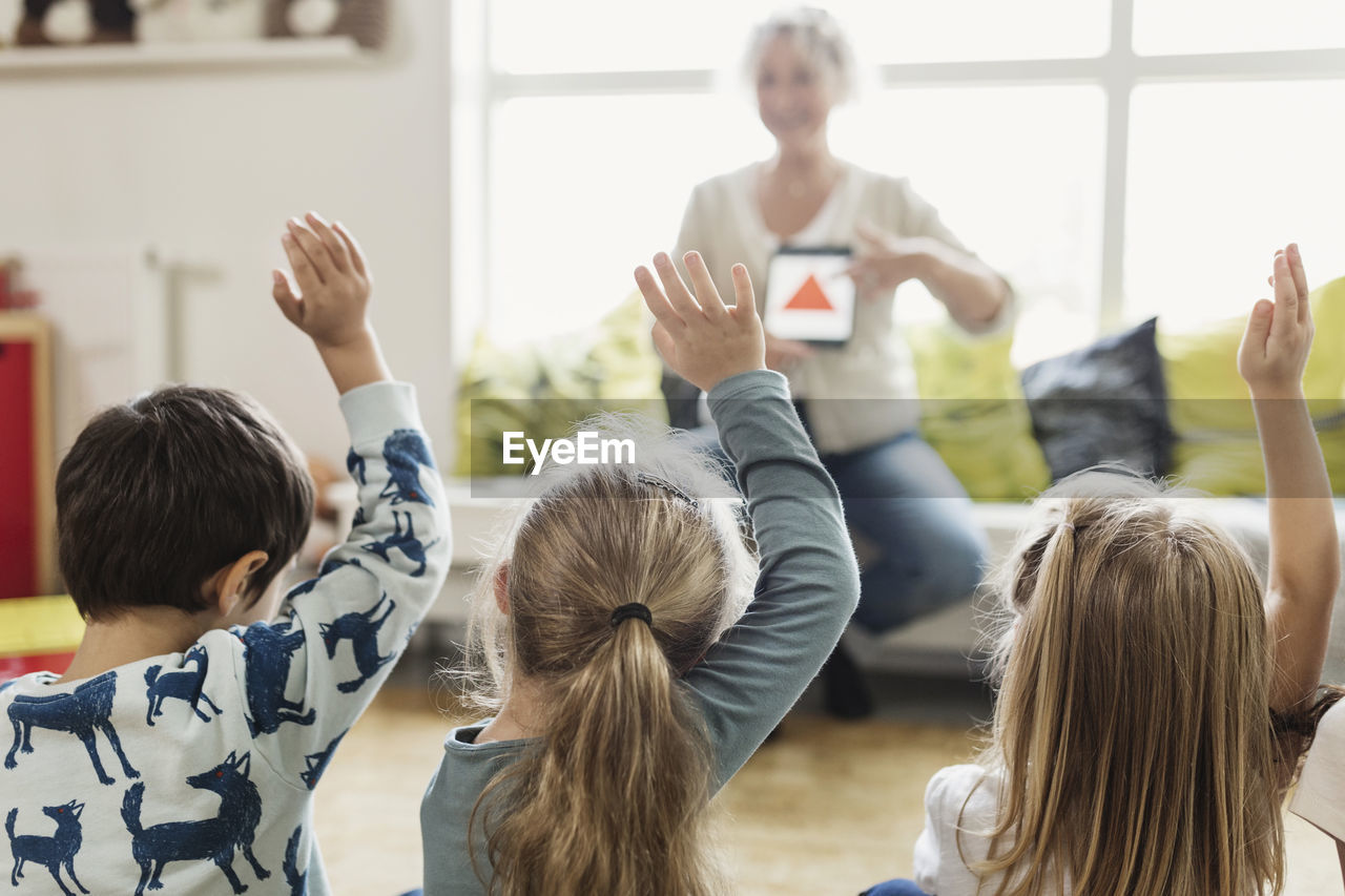 Rear view of students raising hands while teacher showing digital tablet at preschool