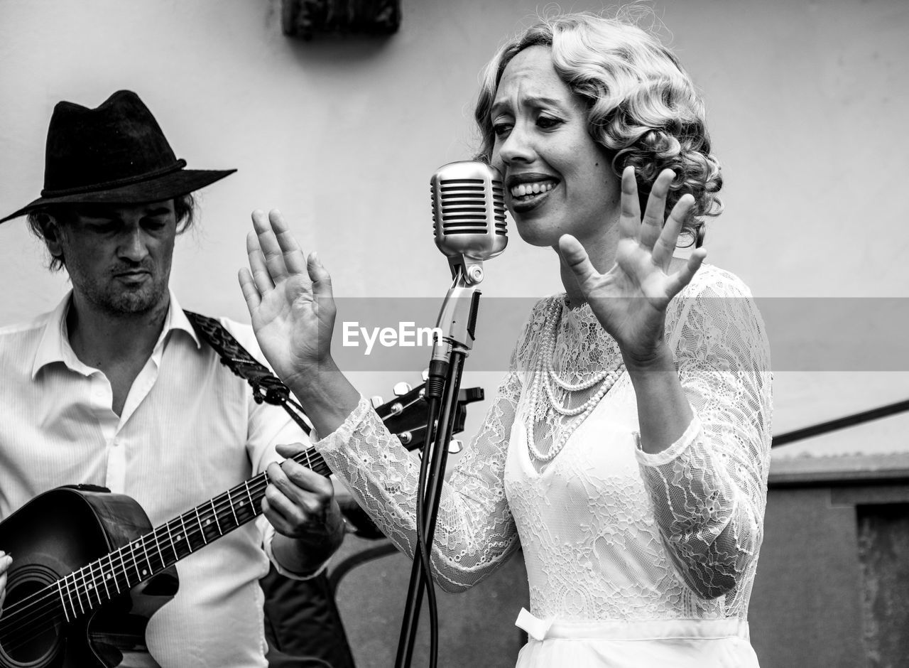 Woman singing on microphone with man playing guitar against wall