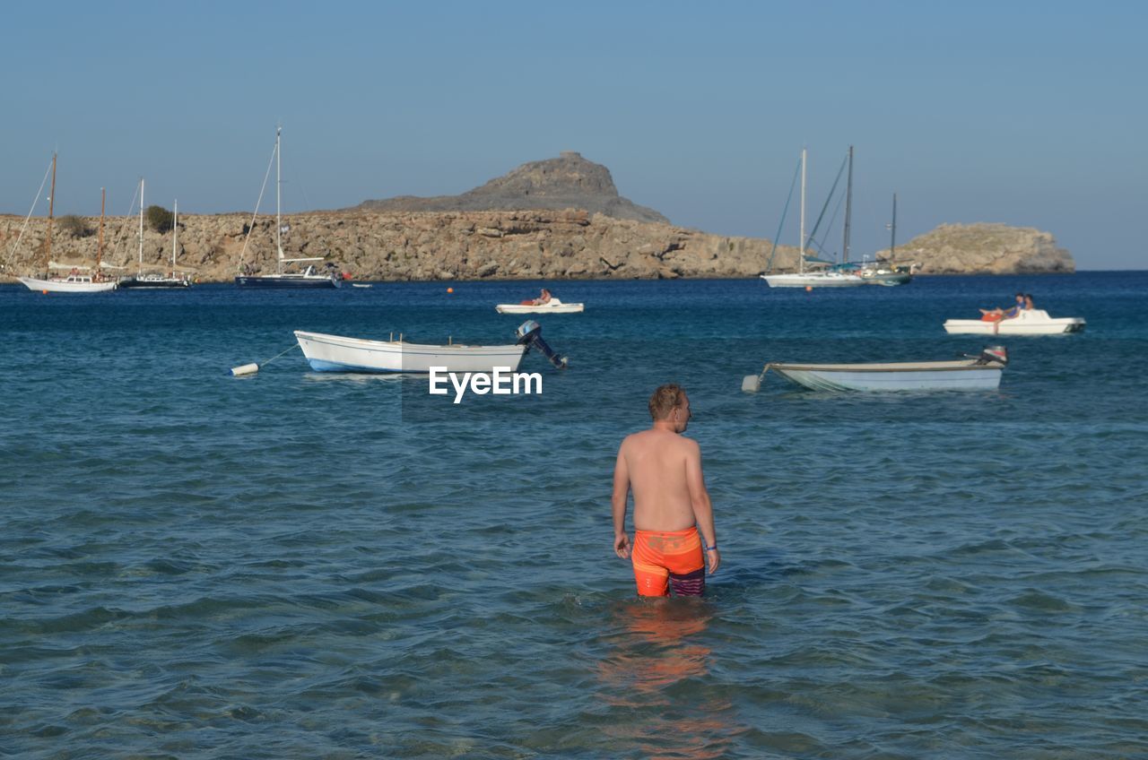 Rear view of shirtless man standing in sea by boats
