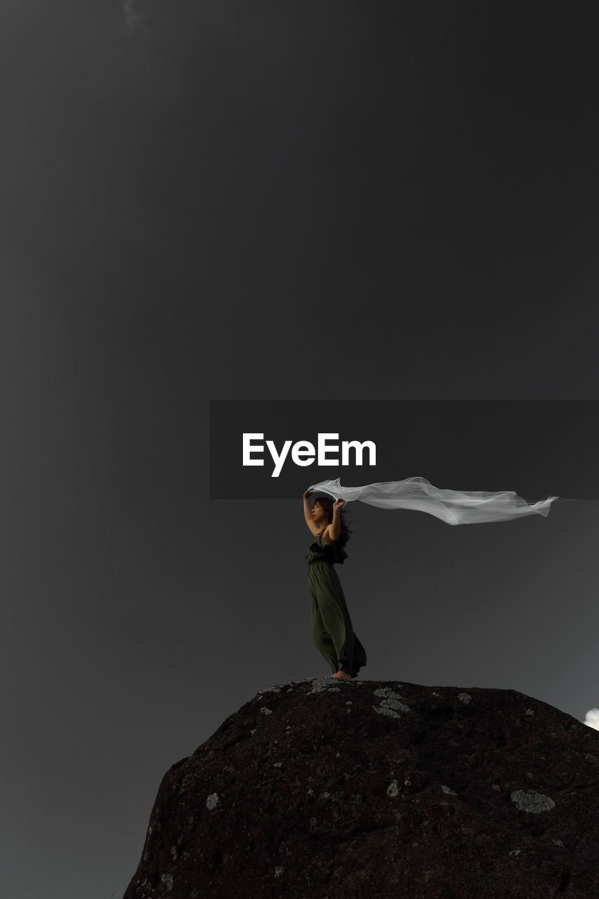 Woman holding scarf while standing on rock formation against sky