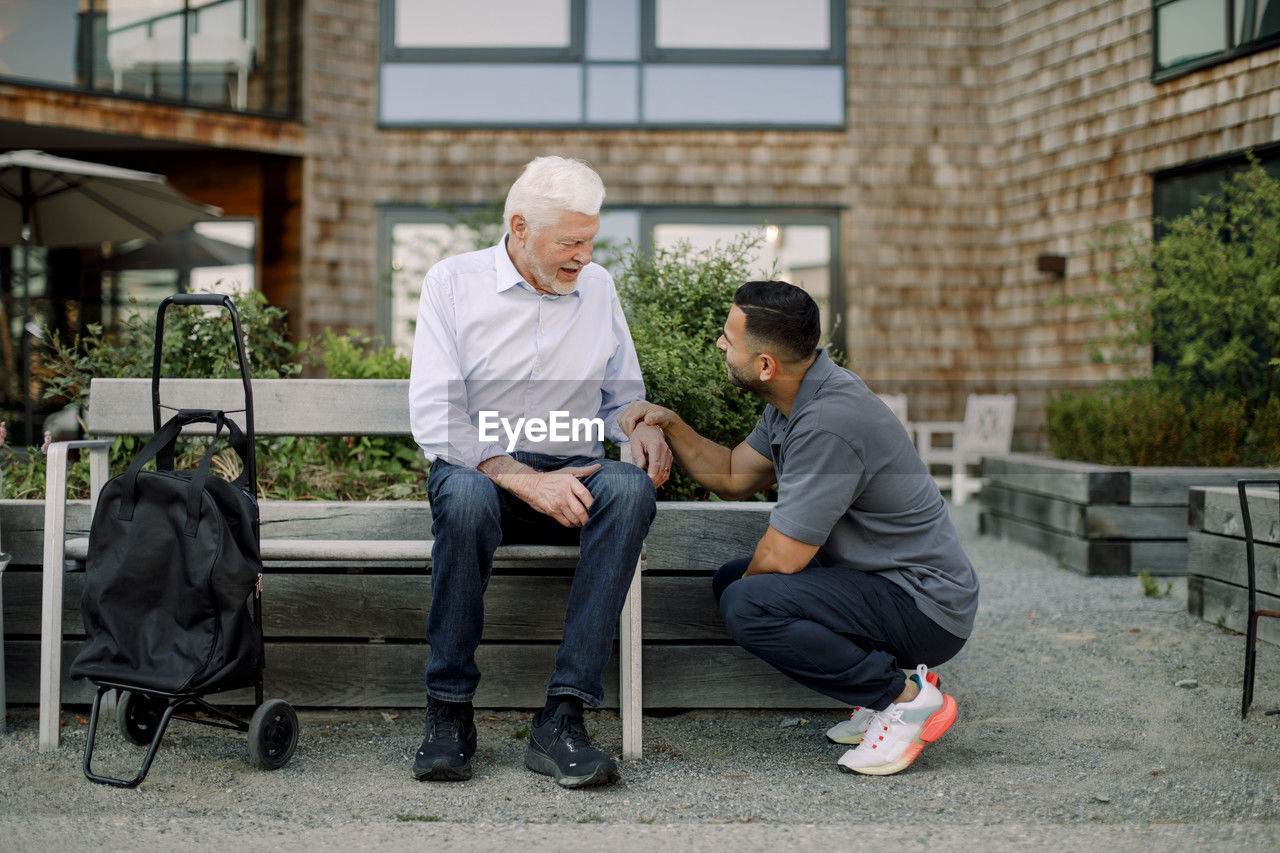 Full length of male nurse squatting while talking to retired senior man sitting on bench at back yard