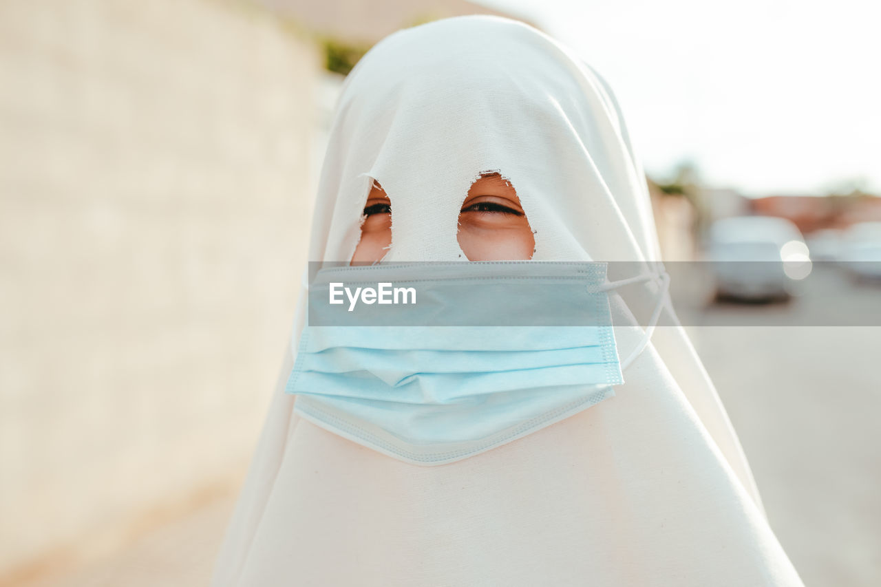 Anonymous spooky person wearing white ghost costume and medical mask standing on street and looking at camera during halloween celebration