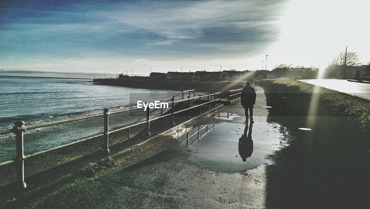 Reflection of person in puddle on footpath by sea against sky