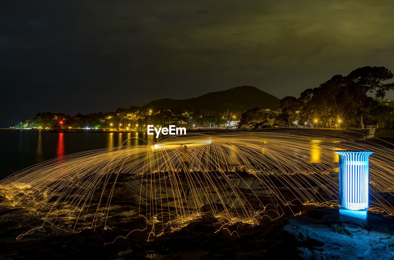 Illuminated light trails by river against sky at night