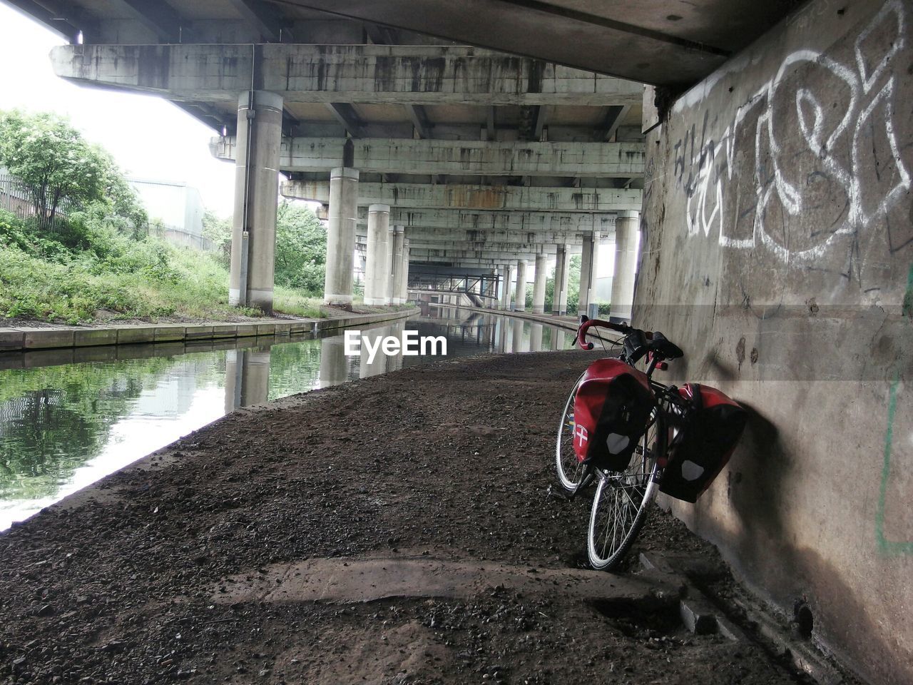 A bicycle stands against a cement wall below a bridge next to a small river