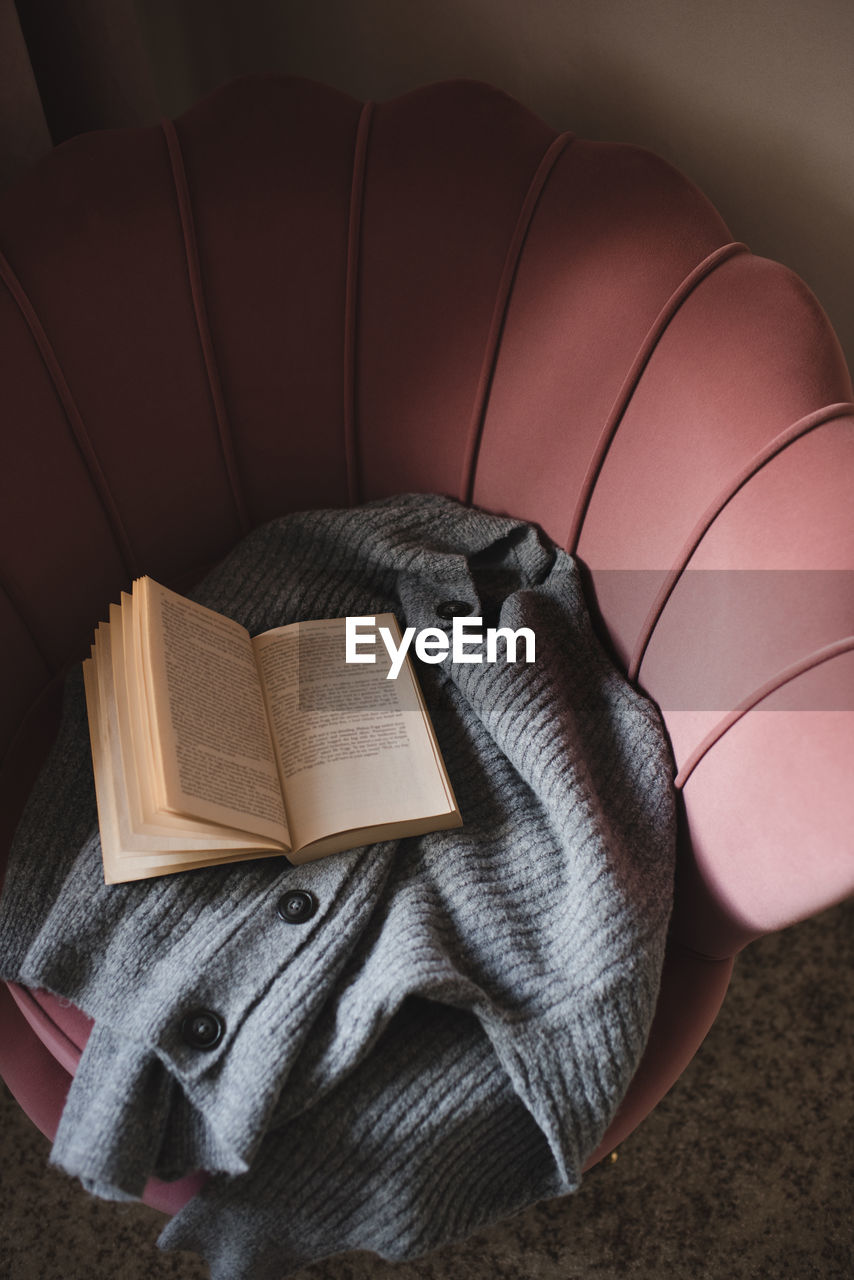 Open paper book with knitted wool fabric on cozy accent chair in room close up.