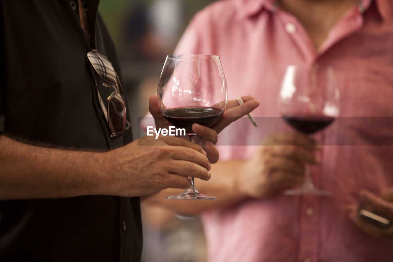 Midsection of men holding wineglasses and cigarettes while standing outdoors