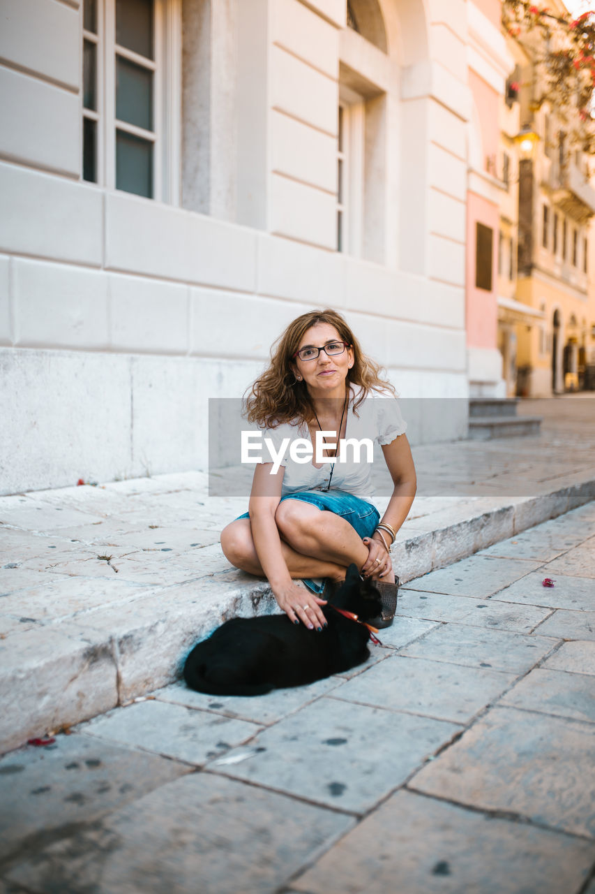 Full length portrait of woman stroking cat on footpath in city