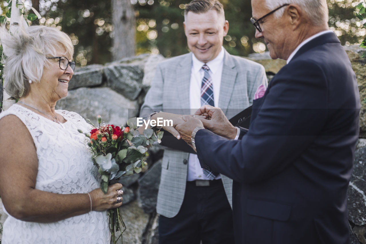 Senior groom putting wedding ring in bride's finger by minister during ceremony