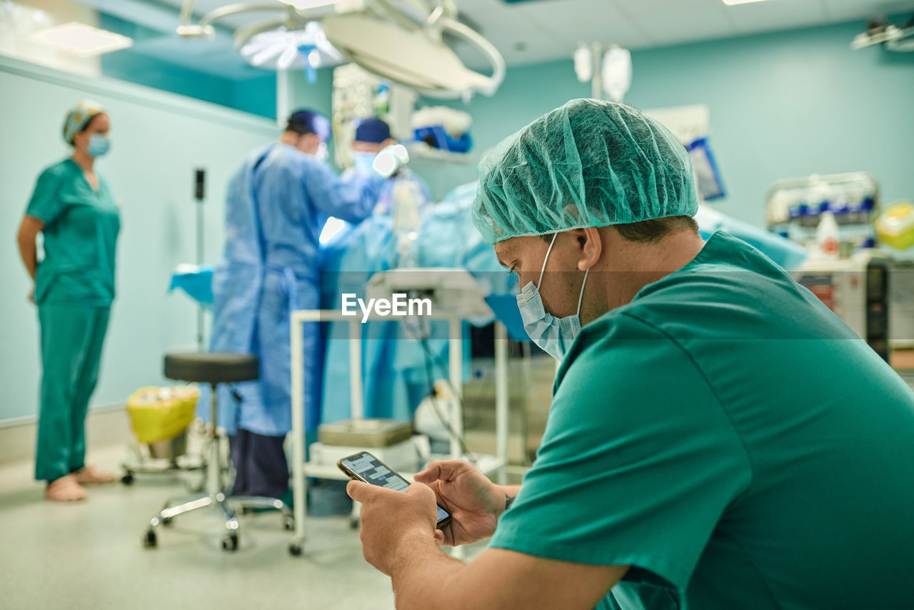 Side view of anonymous concentrated young male assistant in medical uniform and mask messaging on mobile phone while team of professional surgeons performing operation in modern clinic