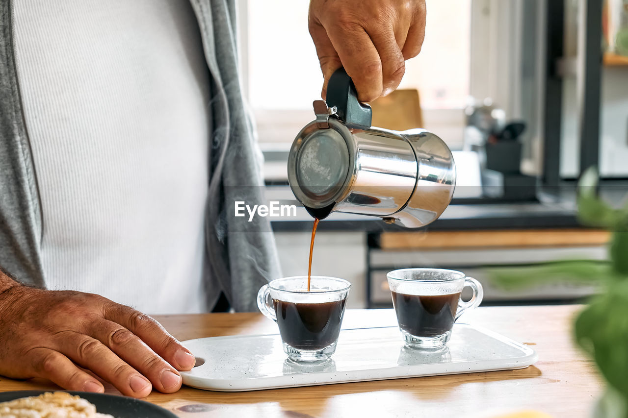 Man preparing classic italian coffee mocha, pouring coffee from moka pot into glass coffee cup.