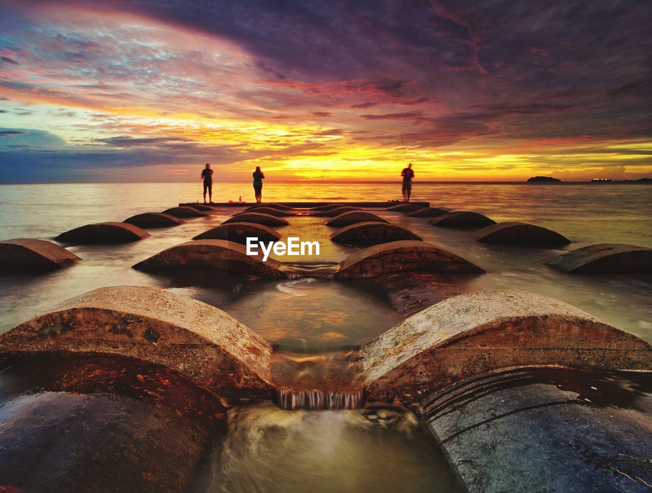 People standing on pipes at beach against sky during sunset