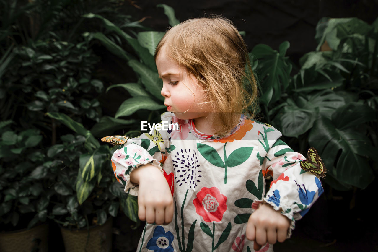 Cute baby girl blowing butterfly on her hand against plants