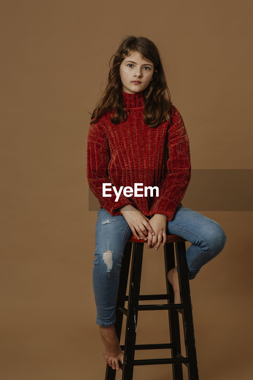 Portrait of serious girl sitting on stool against brown background