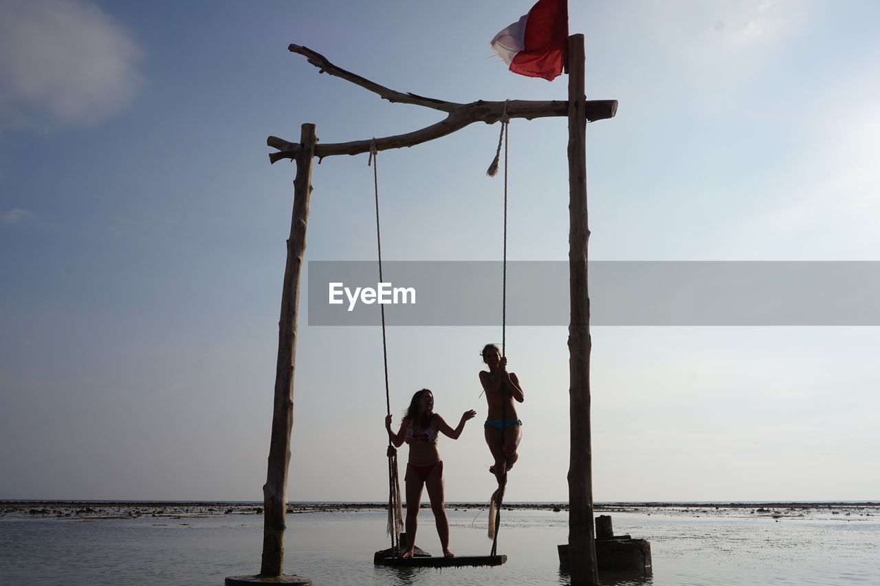 Women standing on swing over sea against sky