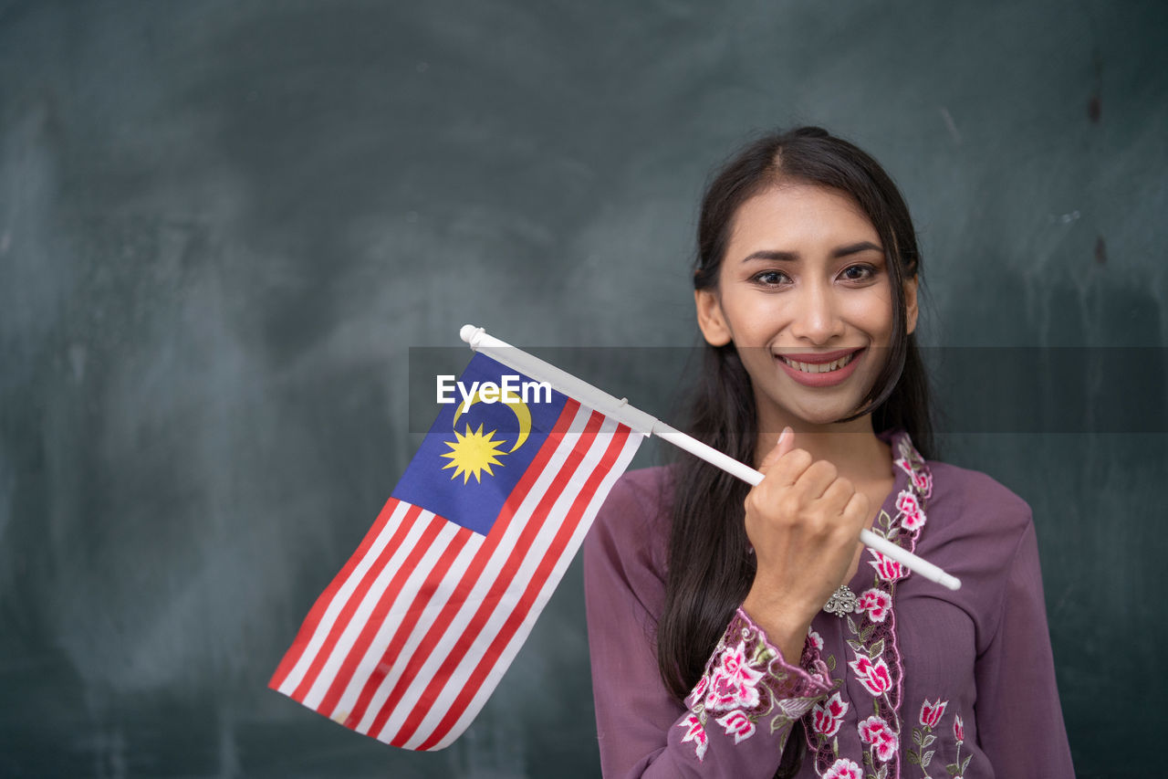 Portrait of smiling young woman holding malaysian flag against blackboard