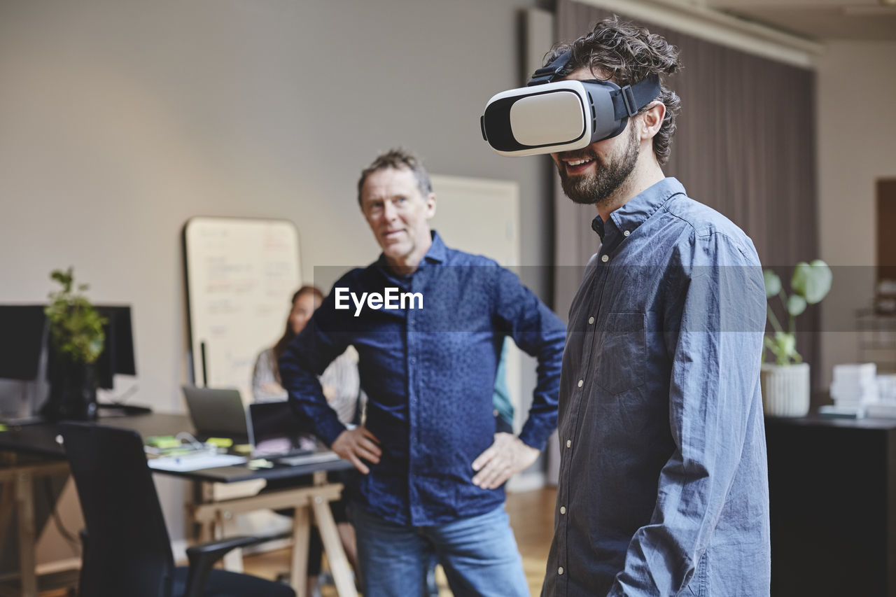 Mature businessman looking at male colleague using virtual reality simulator while standing in creative office