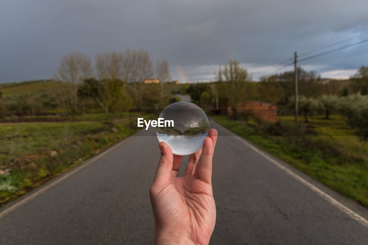 Cropped hand holding crystal ball over road against sky