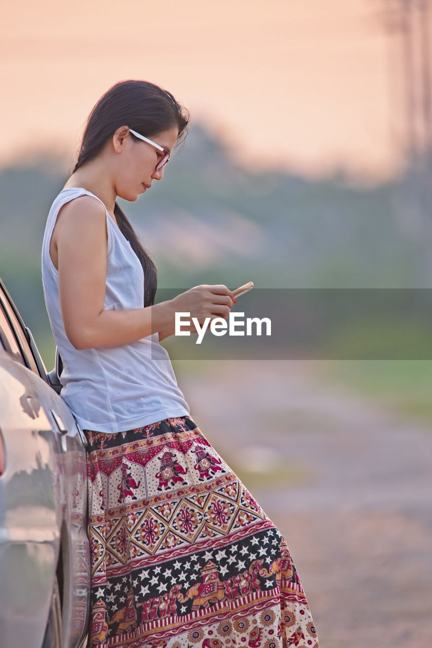 Side view of woman using phone while standing by car during sunset