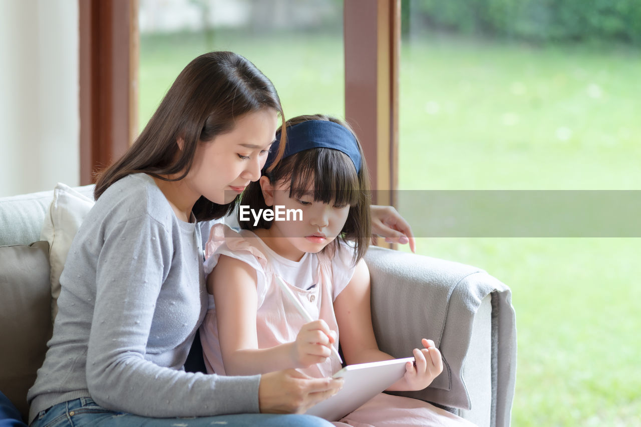 Happy asian mother teaching her cute kid daughter to studying in living room at home