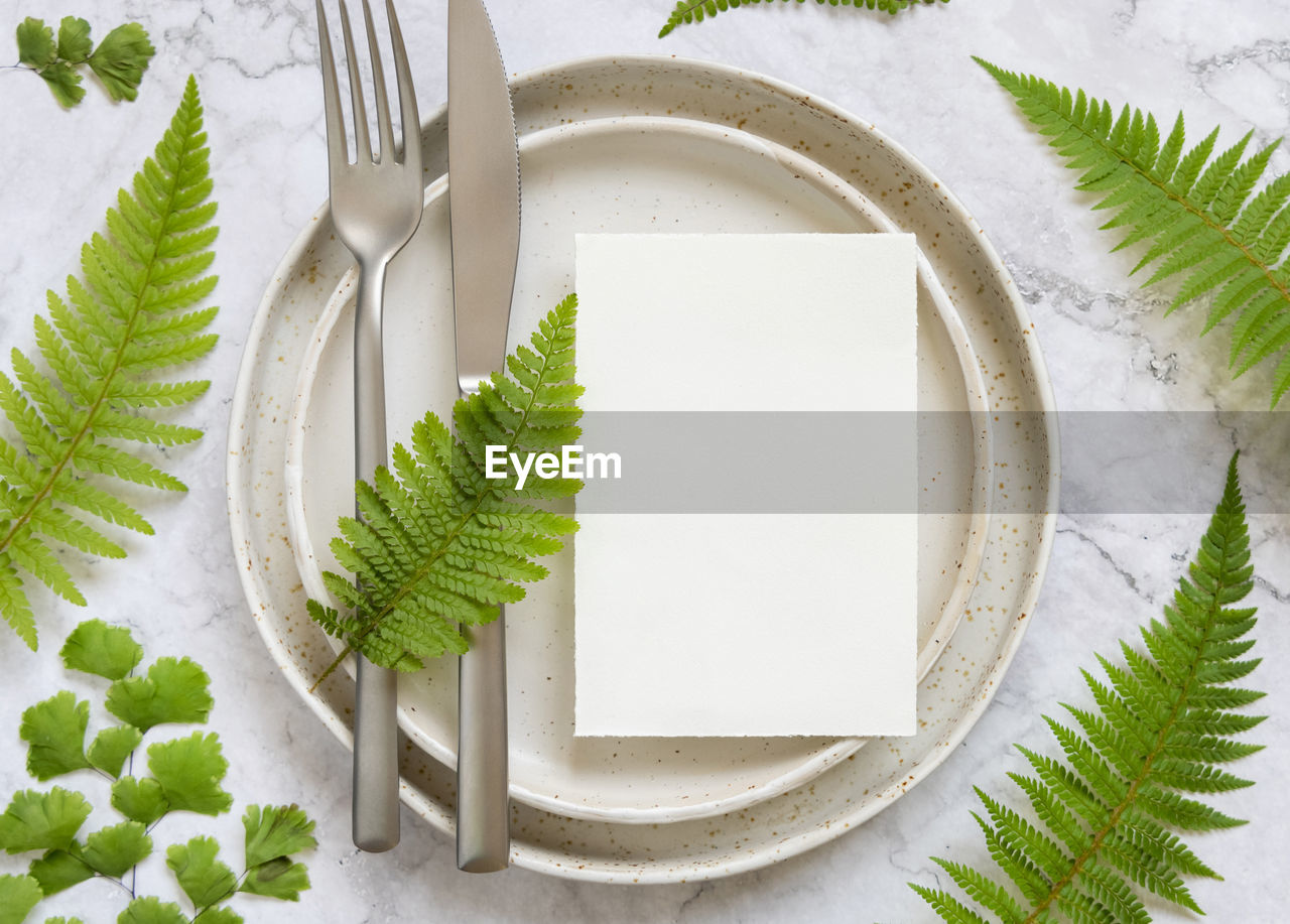 Blank paper card on laying on a plate with fork and knife on marble table with fern leaves 