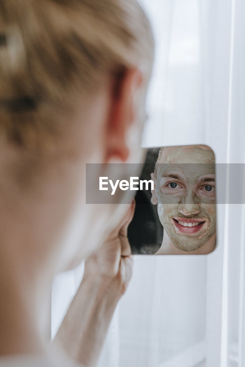 Happy young make-up artist with facial mask looking at mirror