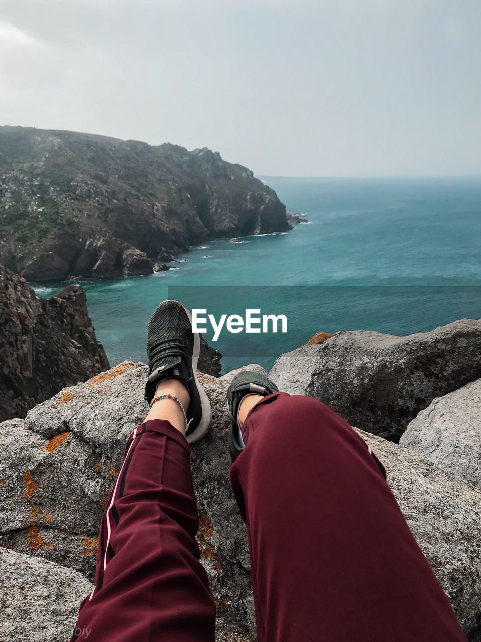 Low section of person relaxing on rocks by sea against sky