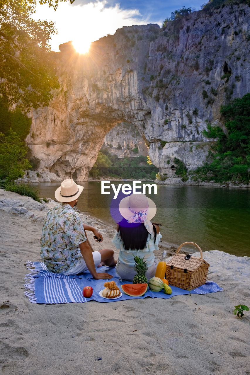 REAR VIEW OF PEOPLE SITTING ON ROCK BY SEA