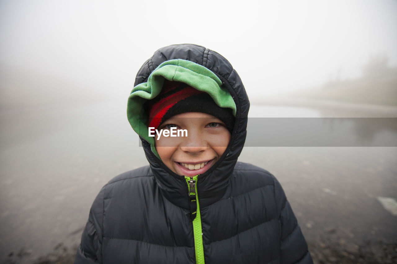 Portrait of smiling boy wearing warm clothing against lake during foggy weather