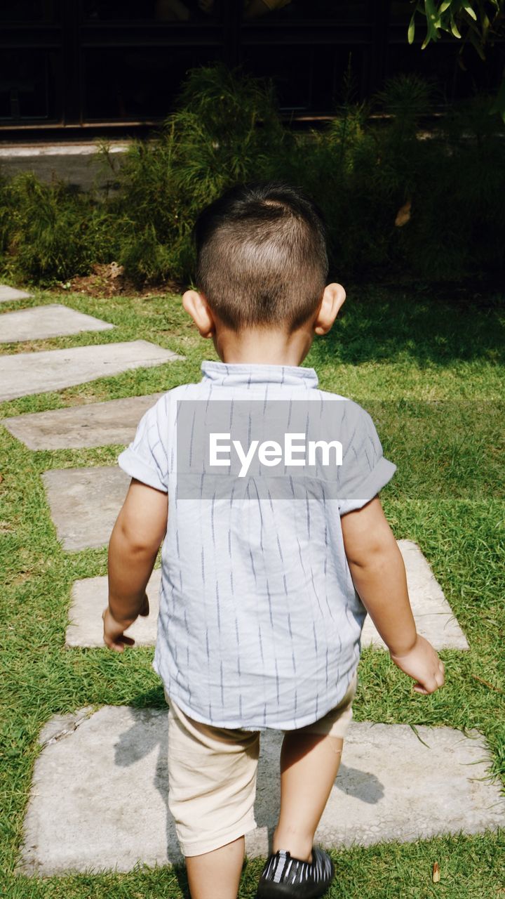REAR VIEW OF BOY STANDING ON FOOTPATH BY PLANTS
