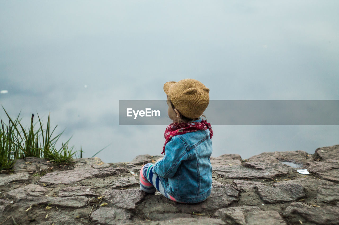 Rear view of boy sitting on rock against cloudy sky