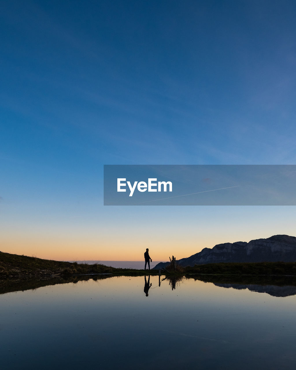 SILHOUETTE MAN ON LAKE AGAINST SKY AT SUNSET