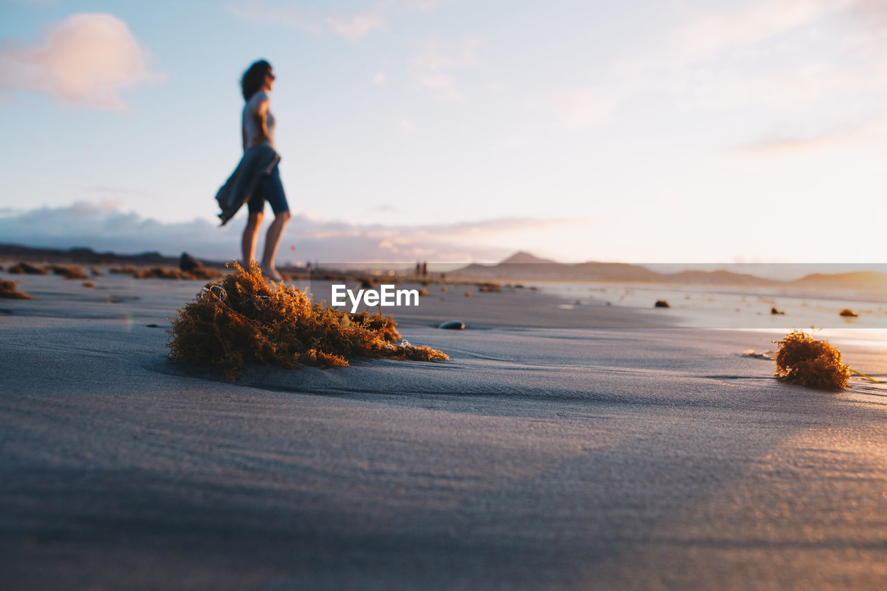sand, land, sky, shore, sea, beach, ocean, water, full length, one person, nature, coast, body of water, morning, adult, holiday, beauty in nature, sunlight, women, horizon, tranquility, vacation, wave, trip, cloud, leisure activity, scenics - nature, young adult, travel, motion, day, side view, solitude, tranquil scene, outdoors, standing, copy space, travel destinations, casual clothing, lifestyles, environment, reflection, walking, idyllic, landscape, relaxation, hairstyle, summer, clothing