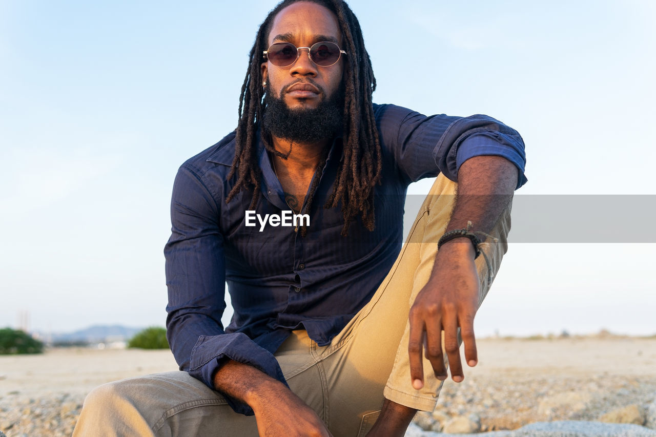 African american man with dreadlocks and sunglasses sitting on sandy shore and looking at camera against cloudless sky in summer