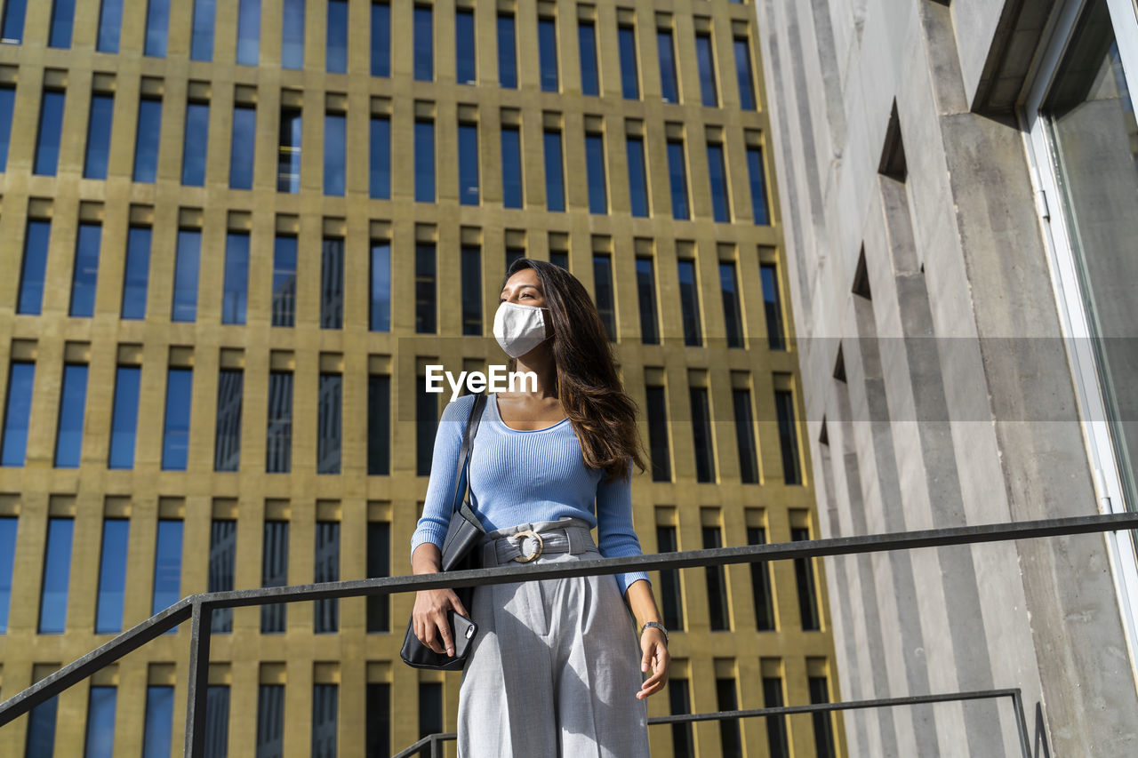 Young businesswoman in face mask looking away while standing against office building during coronavirus outbreak