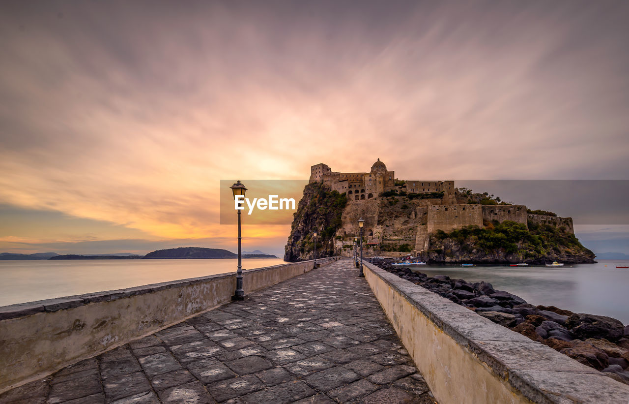 Pier towards castle amidst sea against sky during sunset