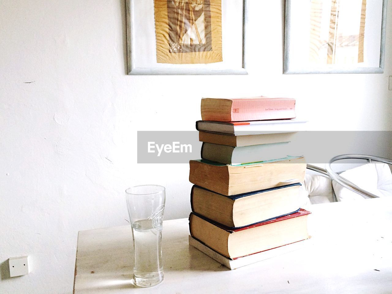 Stack of books with glass of water on table against wall at home