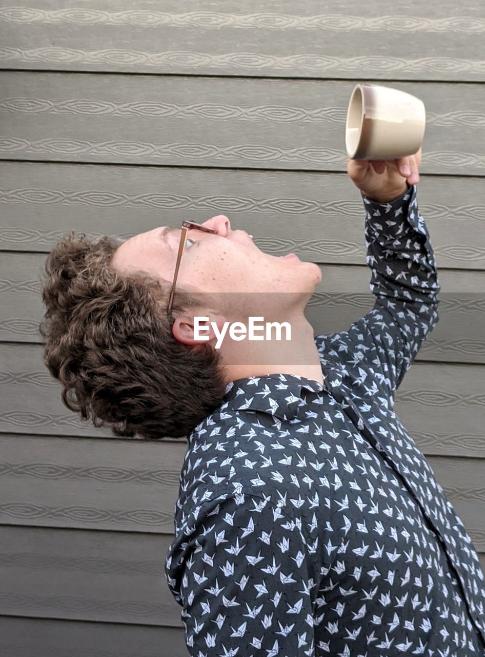  teenage boy with curly hair and glasses standing and  holding a coffee cup above his open mouth