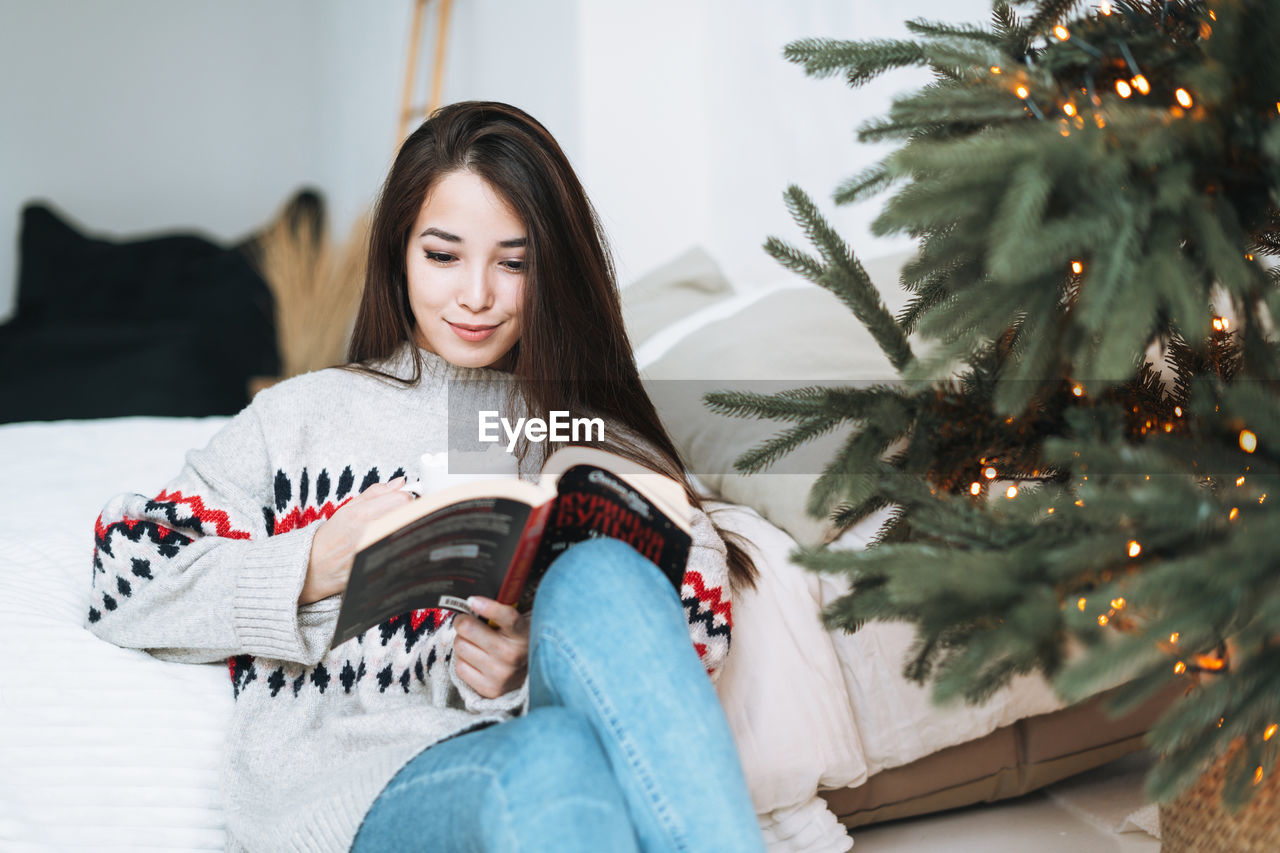 Young asian woman reading book on bed near christmas tree at home