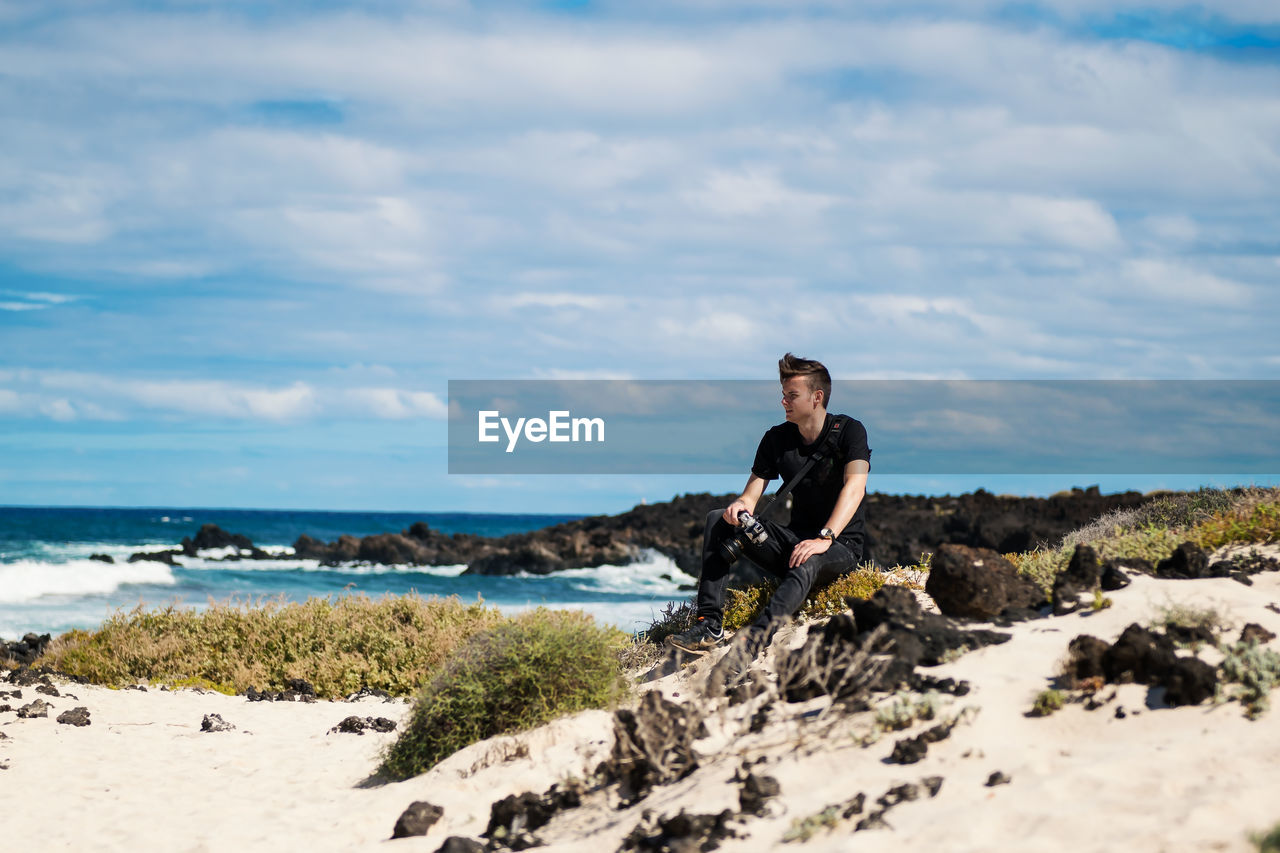 FULL LENGTH OF YOUNG MAN ON ROCK AT BEACH AGAINST SKY