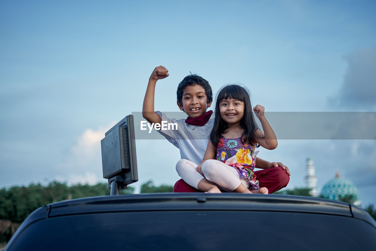 Portrait of smiling siblings sitting on car against sky