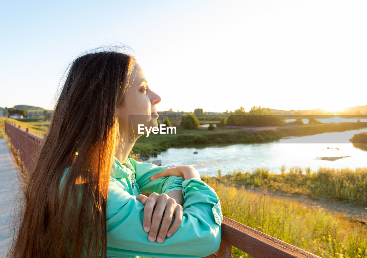 Young girl with loose long hair admiring sunset on river on summer evening. 